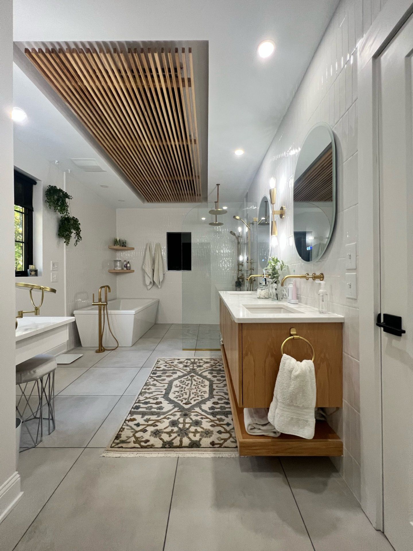 Modern bathroom with wood accents, white walls, and gold fixtures. Features a bathtub, double vanity, and decorative rug.