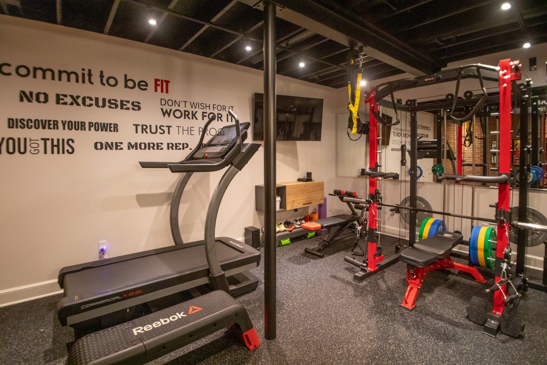 Home gym with treadmill, weight rack, and inspirational wall art. Black and red equipment on a rubber floor.