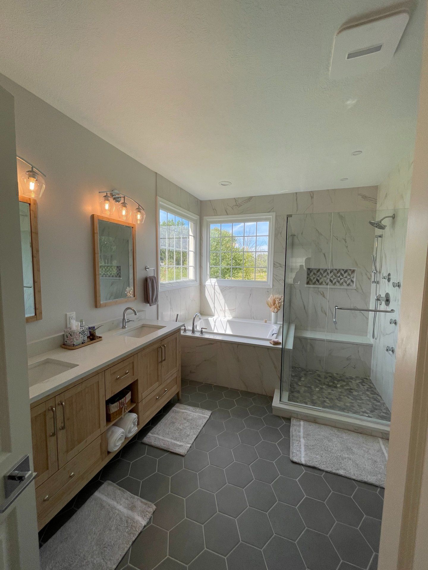 Bathroom with dual sinks, shower, tub, and windows. Gray hexagonal floor tiles and light wooden vanity.