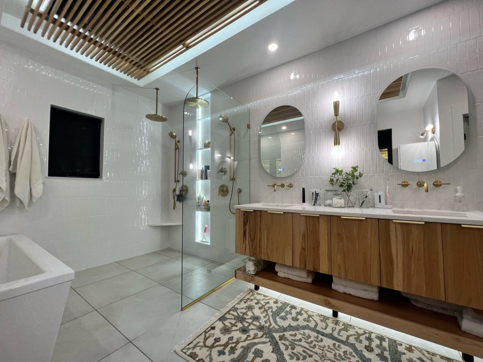 Modern bathroom with wood vanity, glass shower, and round mirrors. White tile, gold fixtures.