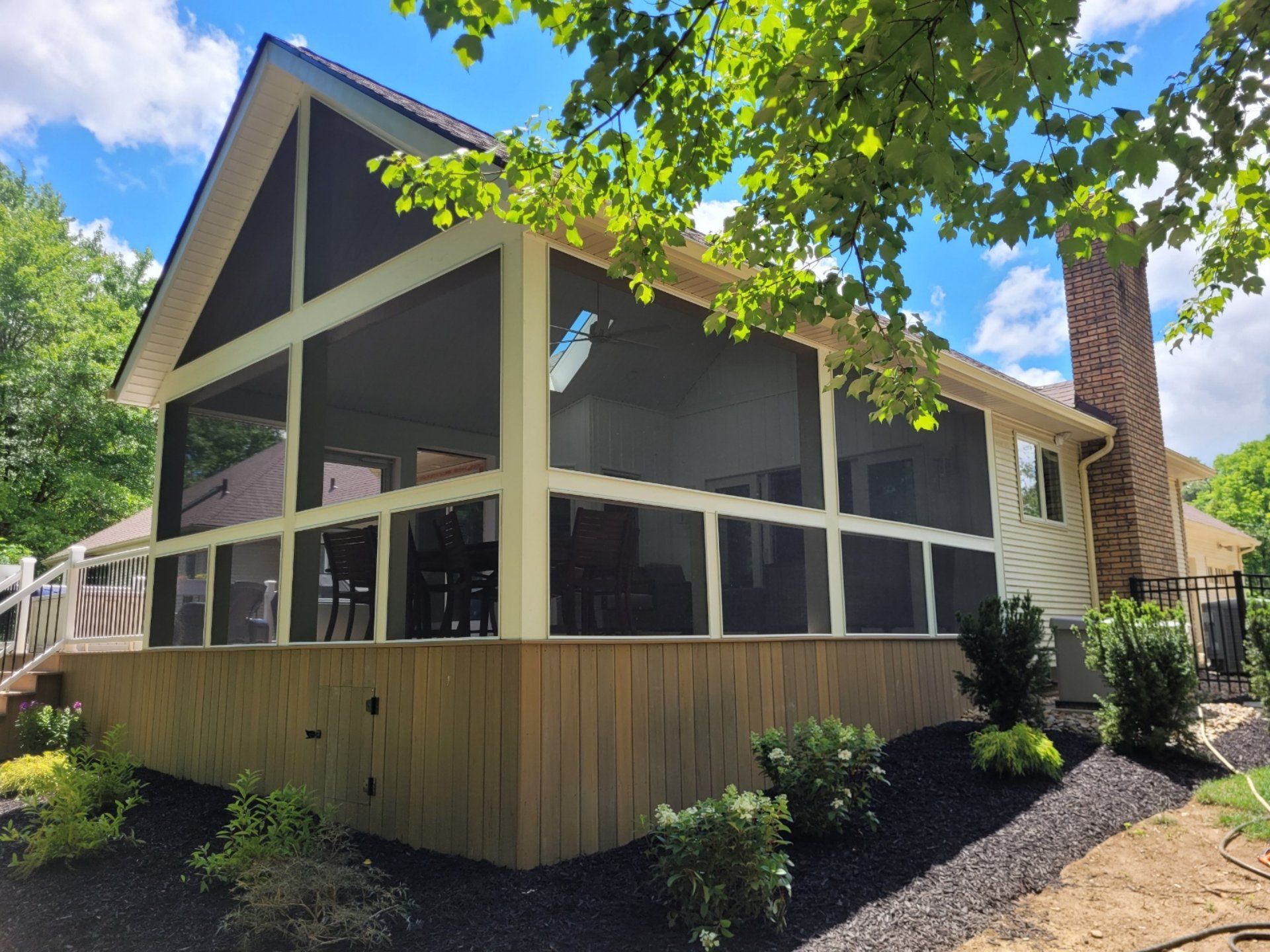 Screened porch with light brown wood siding, dark screens, and a chimney. Blue sky peeks through the trees.