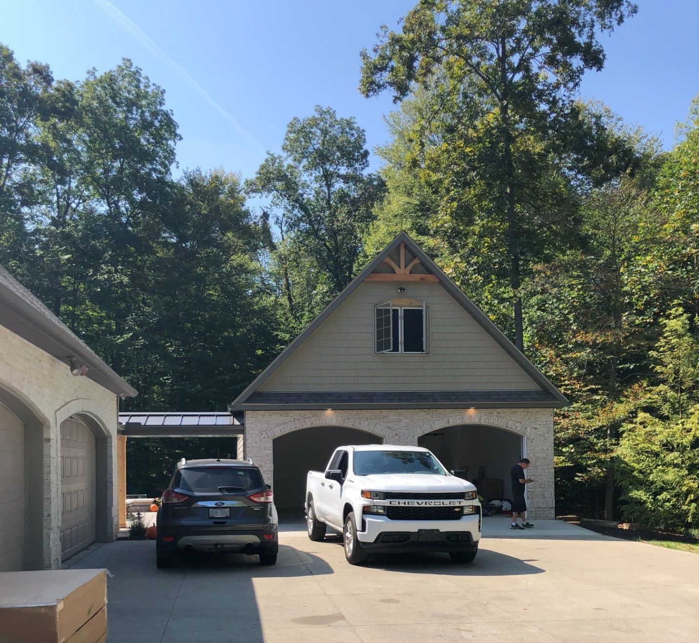 Two vehicles parked in front of a tan and brick garage with trees in the background under a blue sky.