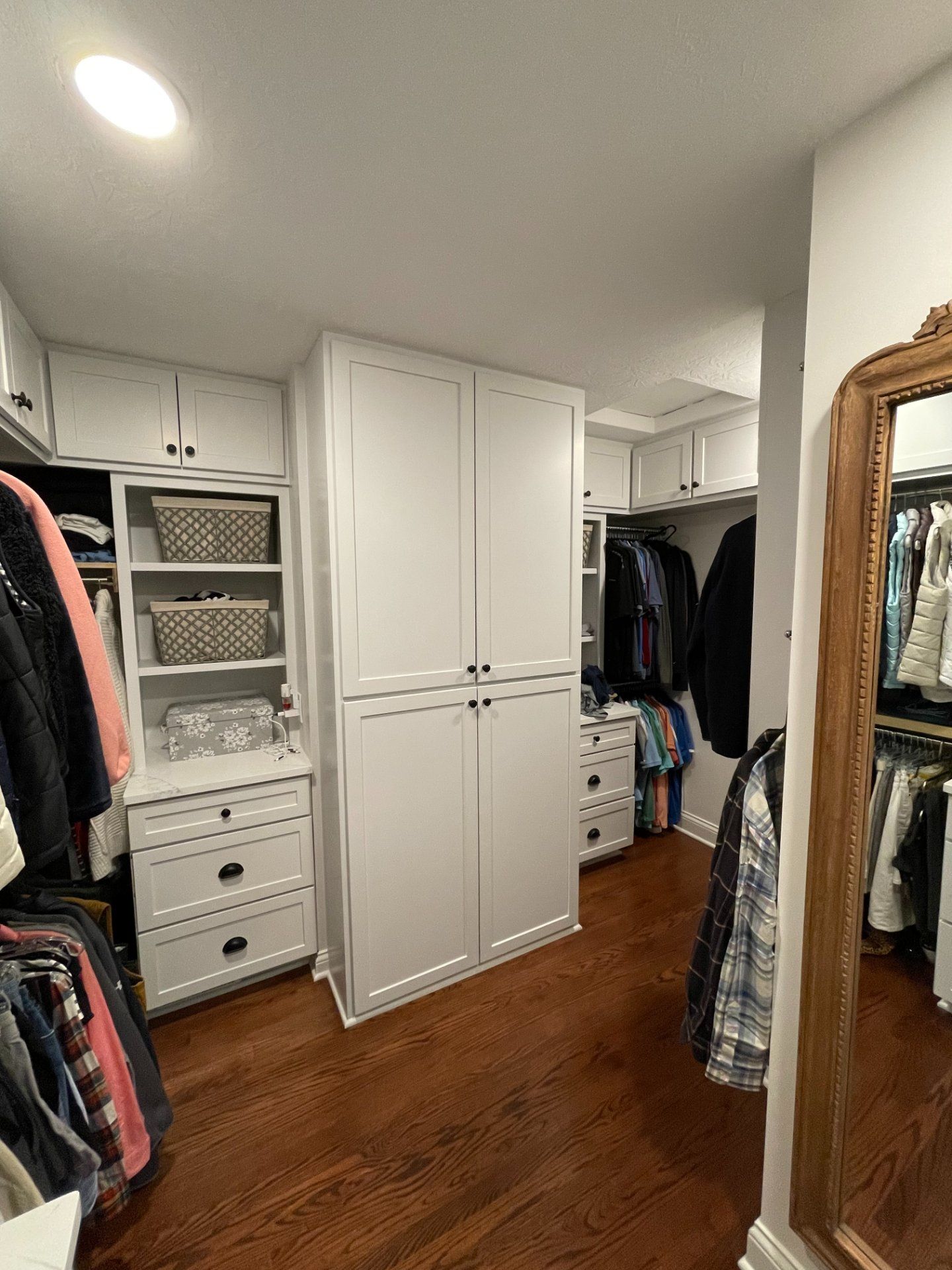 Walk-in closet with white cabinets, wood floors, clothes hanging, and a large mirror.