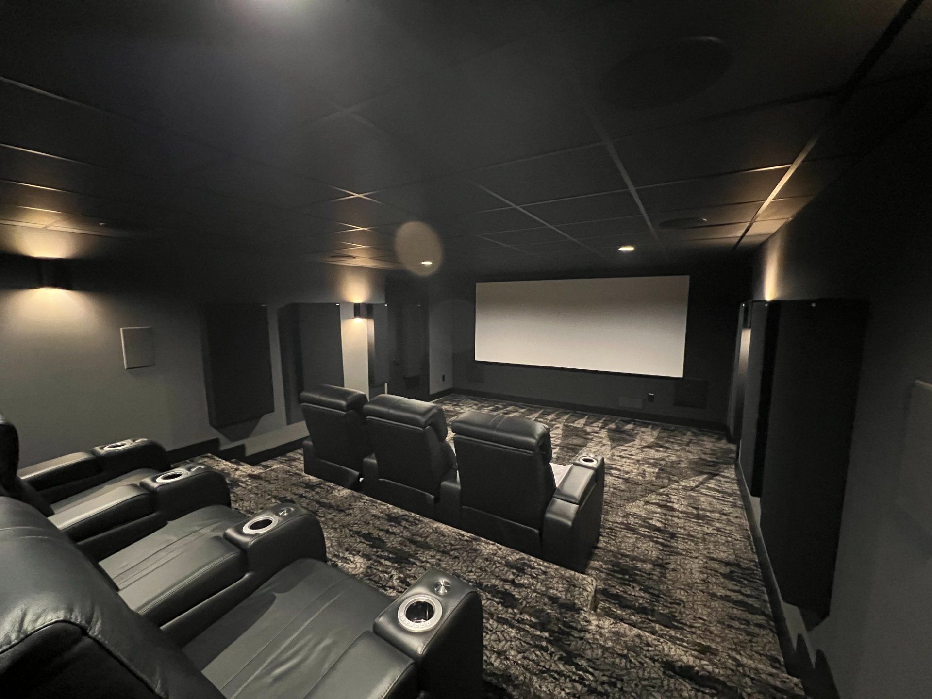 Home theater with black leather recliners, screen, speakers, and patterned carpet. Dark room with recessed lighting.