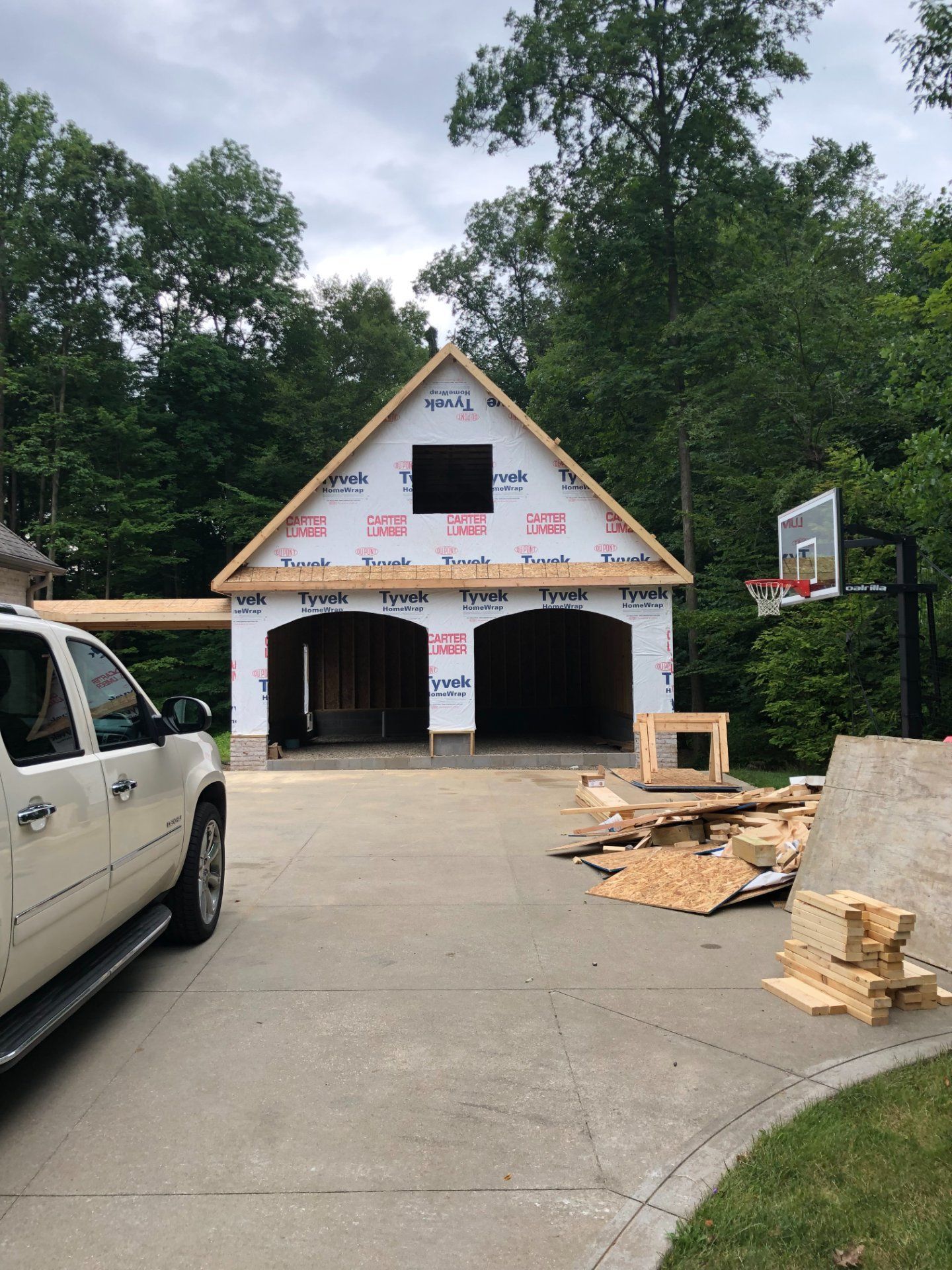 Garage under construction with two bays, white truck, basketball hoop, and trees.