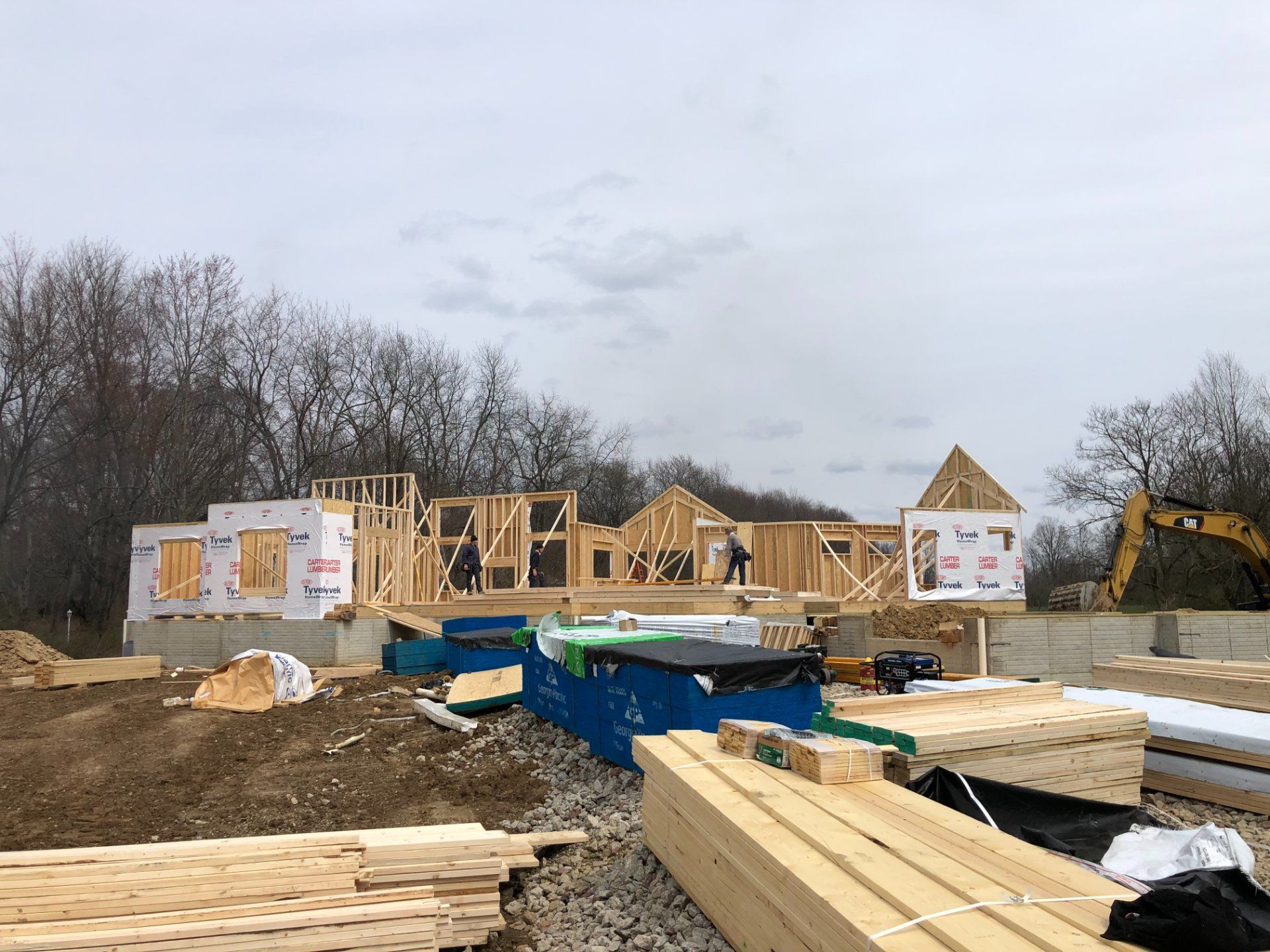 Construction site of a house; wooden frame, materials and equipment in front, two workers visible.
