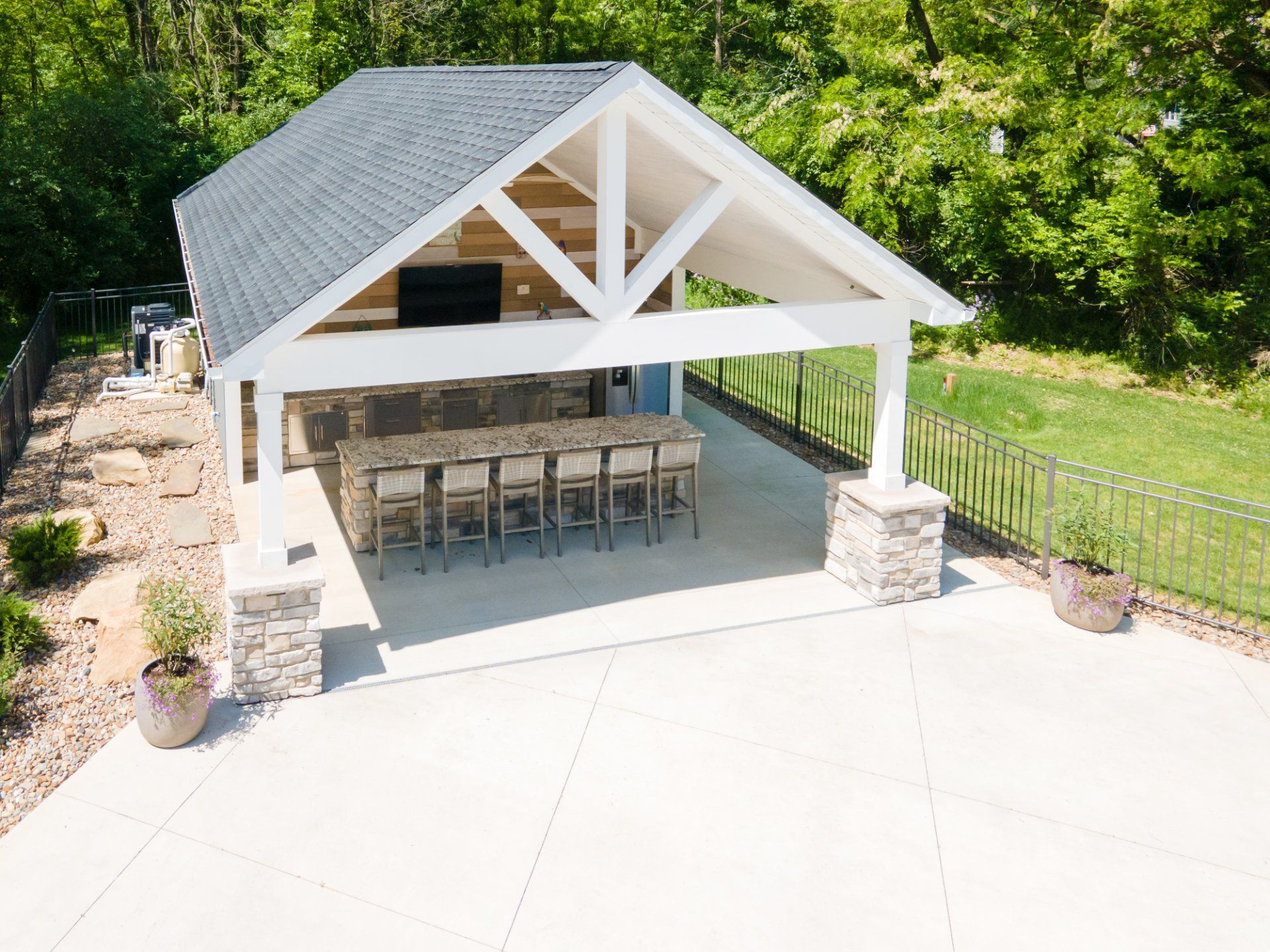 Outdoor covered patio with bar seating, gray roof, stone accents, and surrounding greenery.