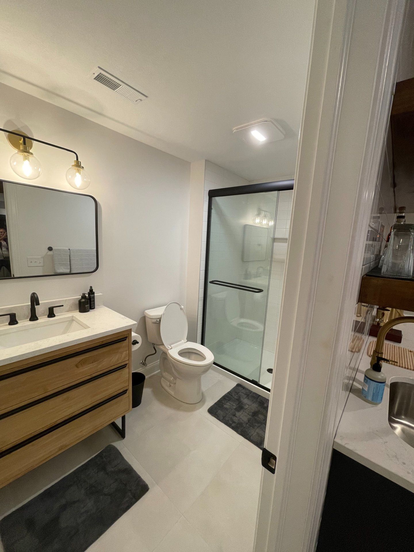 Bathroom with light wood vanity, black framed mirror, and glass shower door. White walls, gray floor mats.