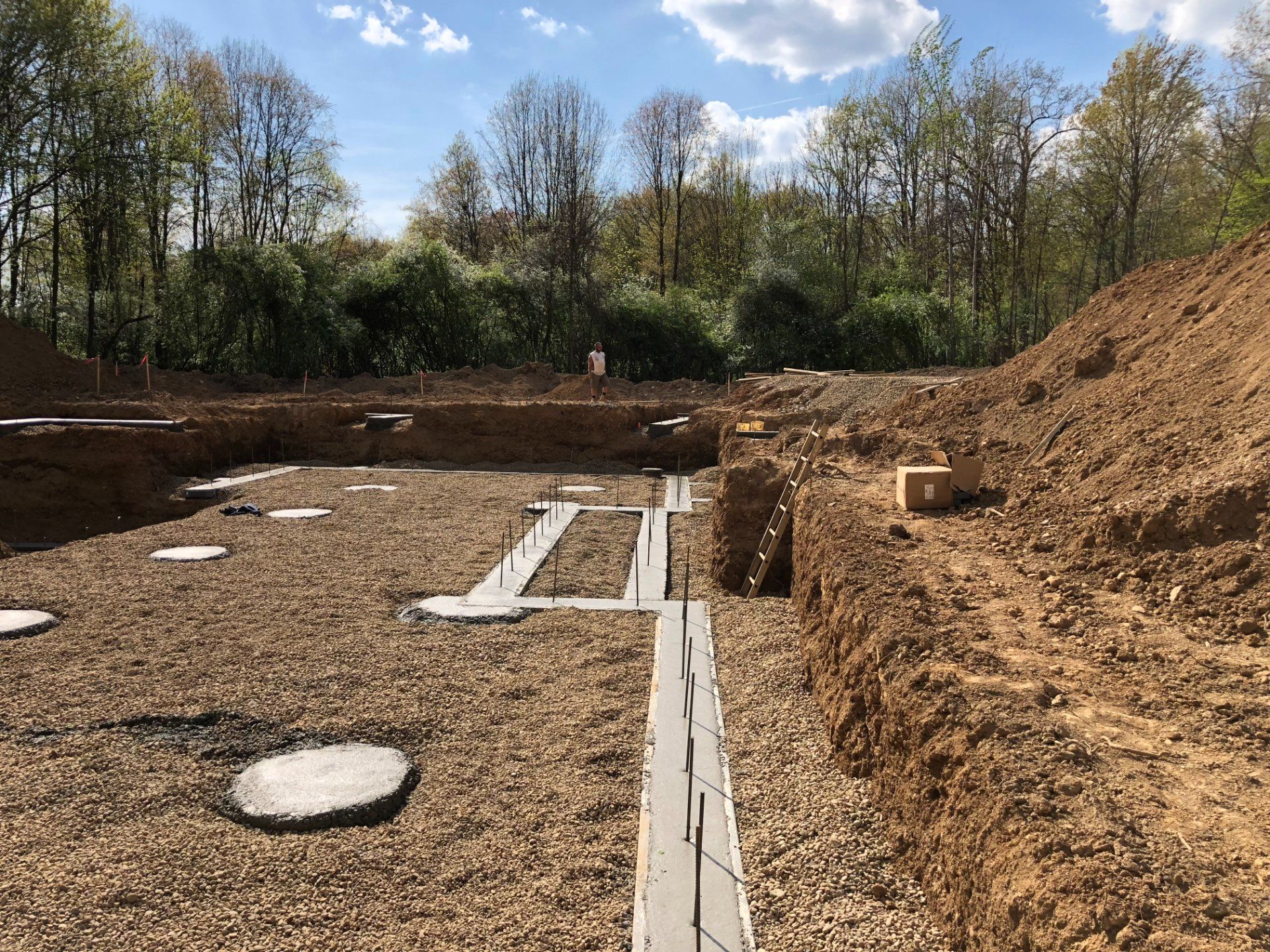 Construction site: foundation excavation with concrete forms and gravel base, trees in background, sunny day.