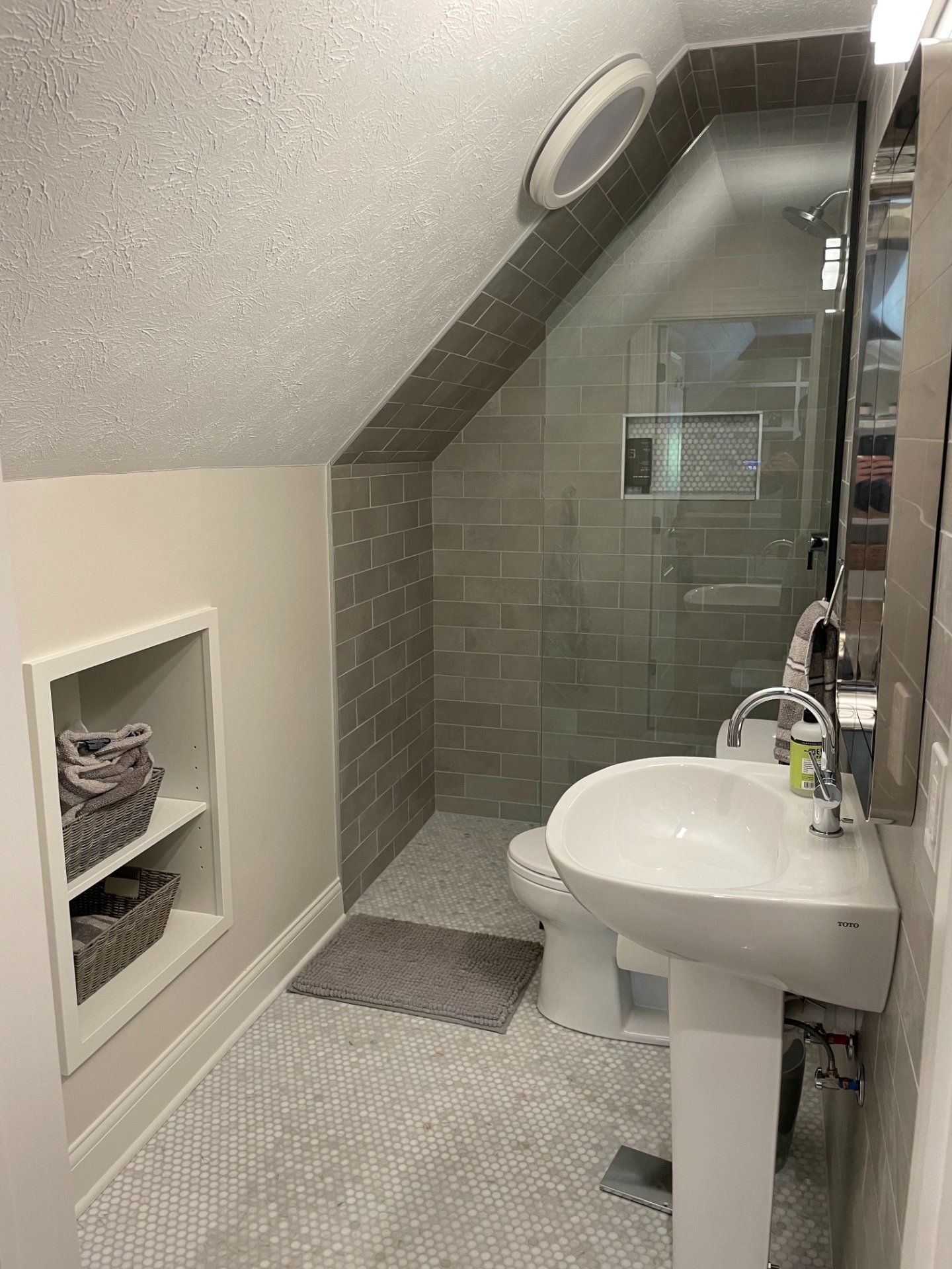 Small bathroom with slanted ceiling, gray tile shower, pedestal sink, and built-in shelf.