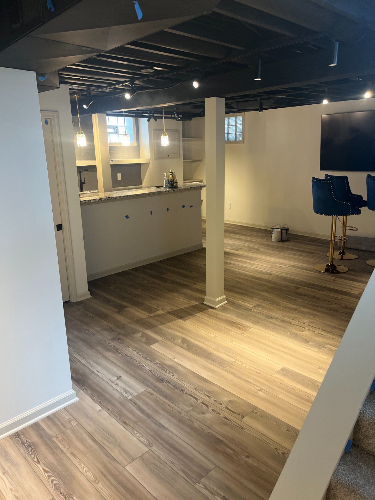 Basement interior with bar, stools, and exposed ceiling beams. Wood-look flooring. White walls and columns.