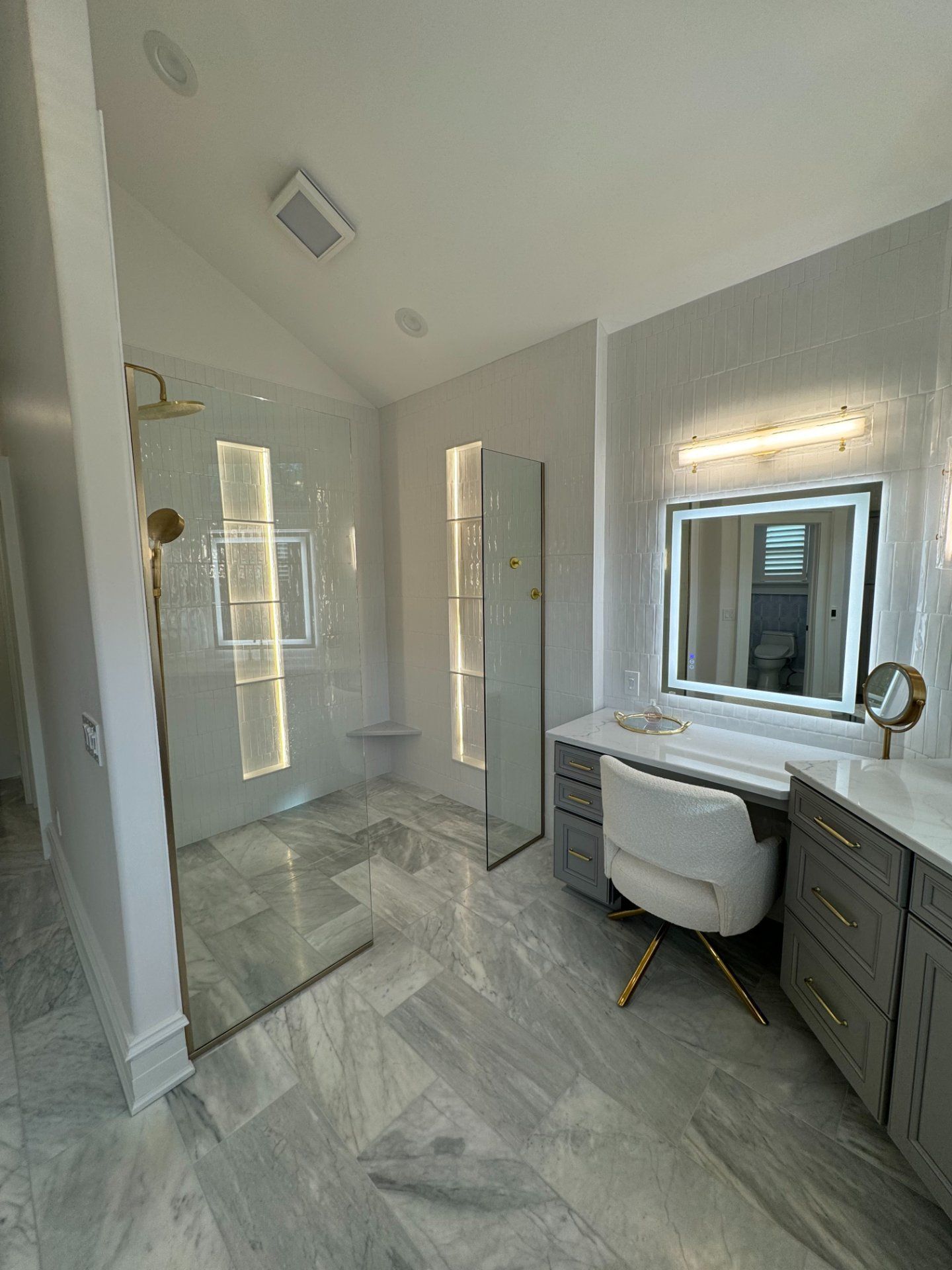 Luxurious bathroom with glass shower, makeup vanity with illuminated mirror, and gray cabinetry.