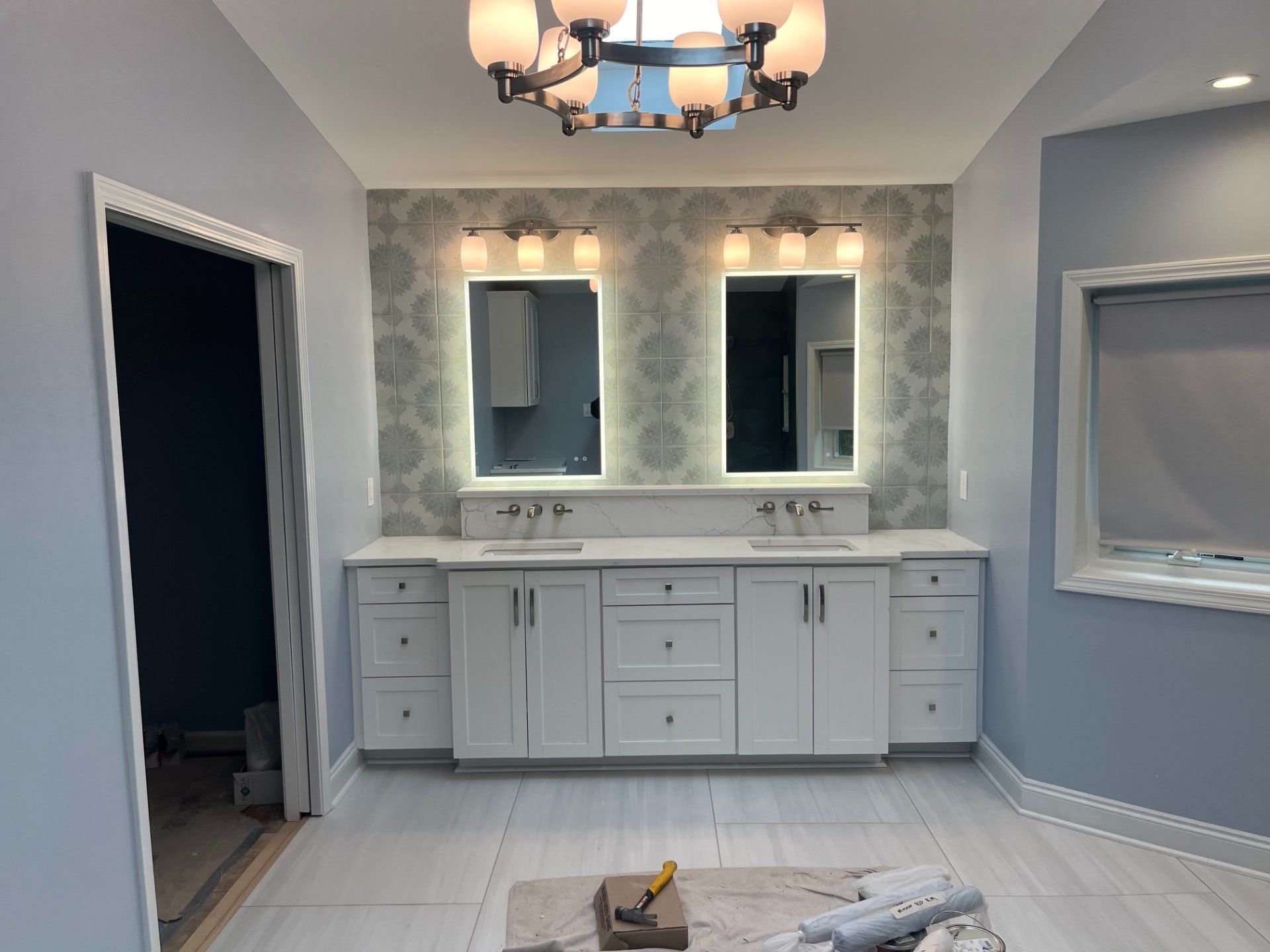 Bathroom with white cabinets, two illuminated mirrors, and patterned wallpaper.