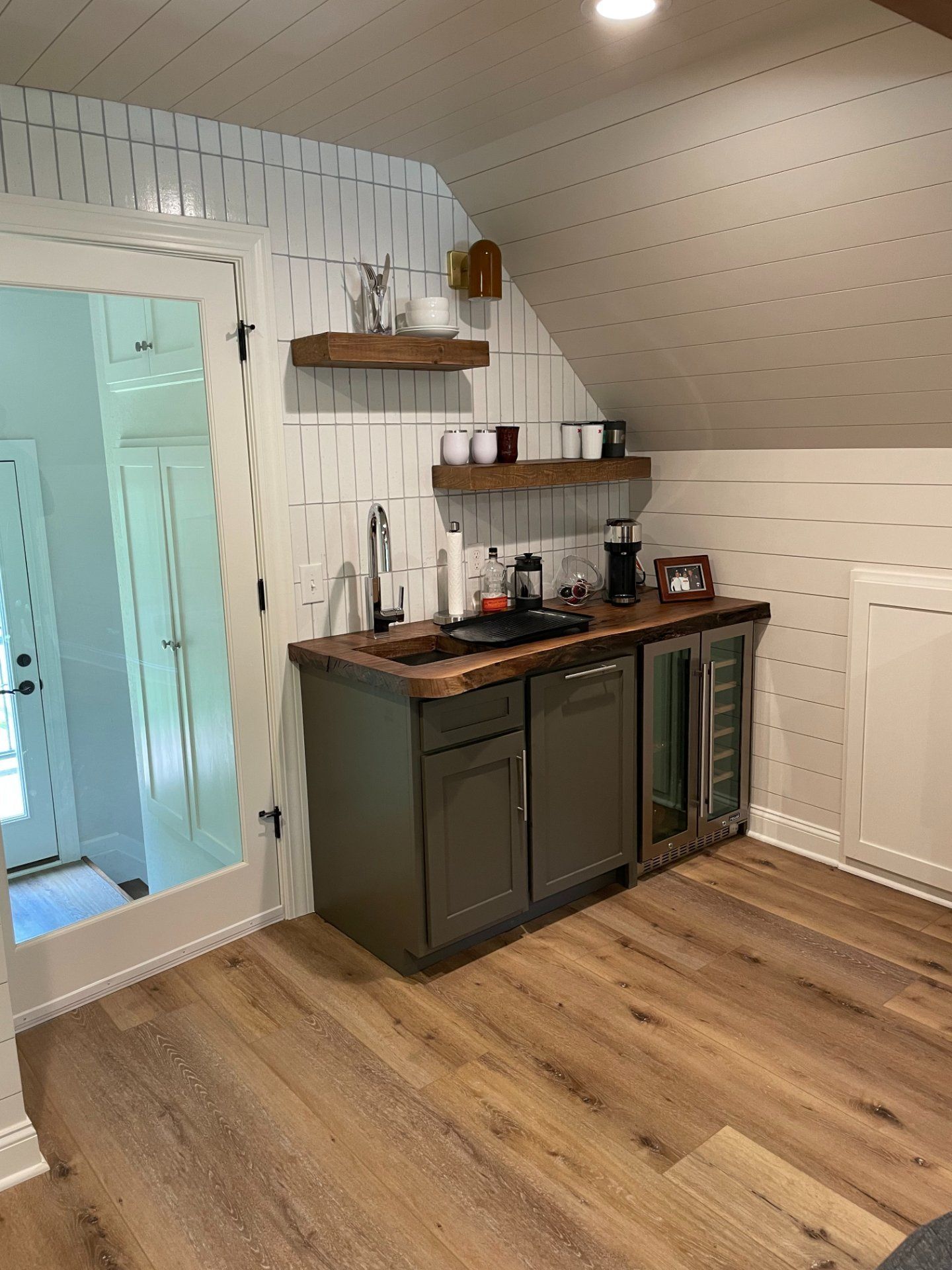 Small kitchen area with a sink, mini-fridge, and cabinets in an angled attic room with wood shelves and flooring.
