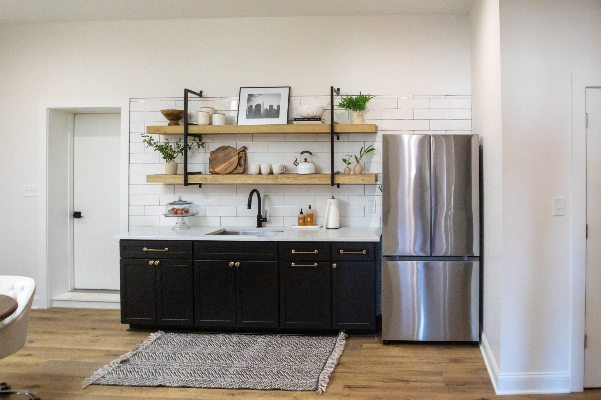 Kitchen with black cabinets, open shelving, stainless steel fridge, white shiplap, and a rug on wooden floors.