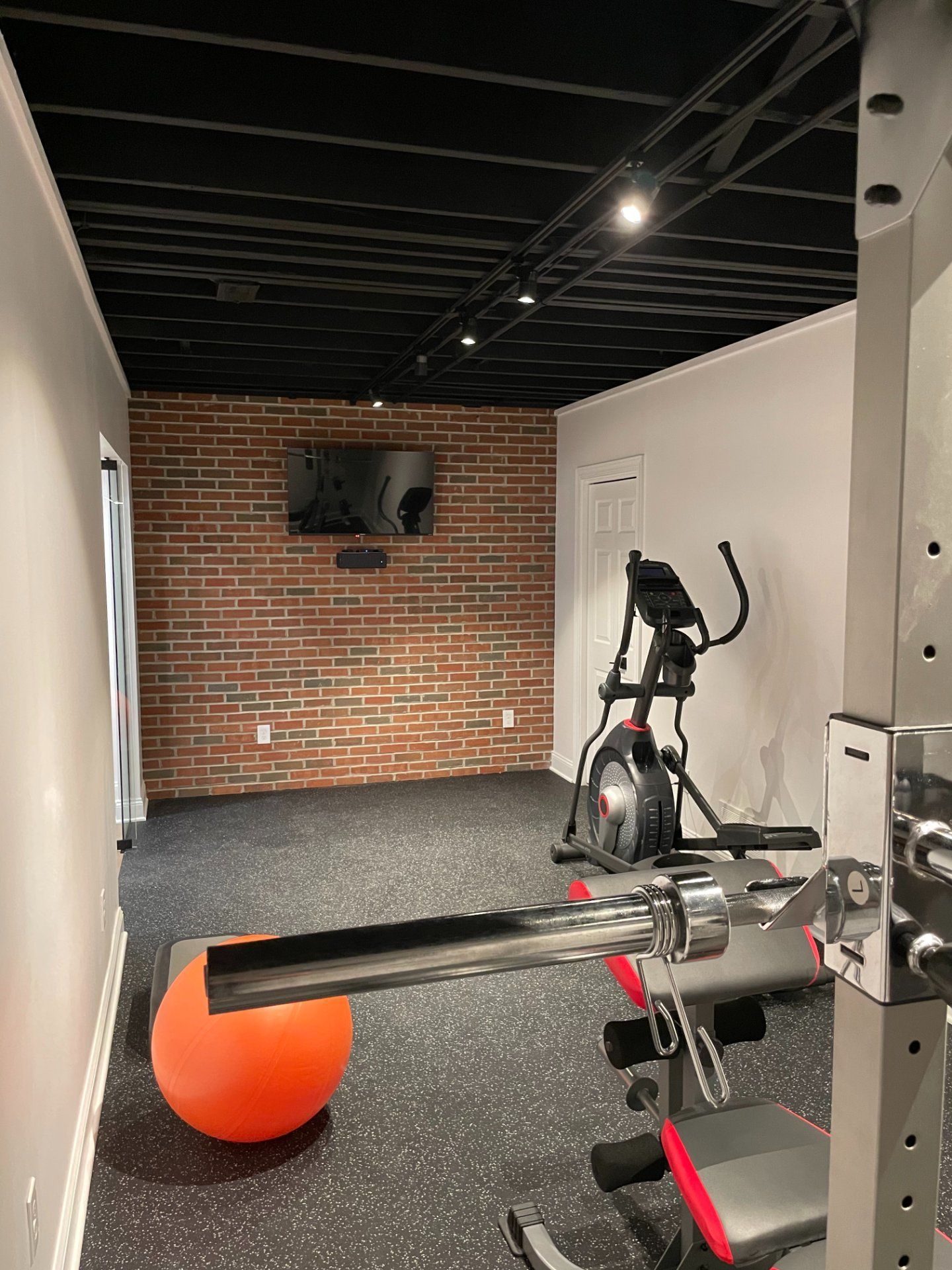 Home gym with exercise equipment: stationary bike, weight machine, and orange exercise ball. Brick accent wall.