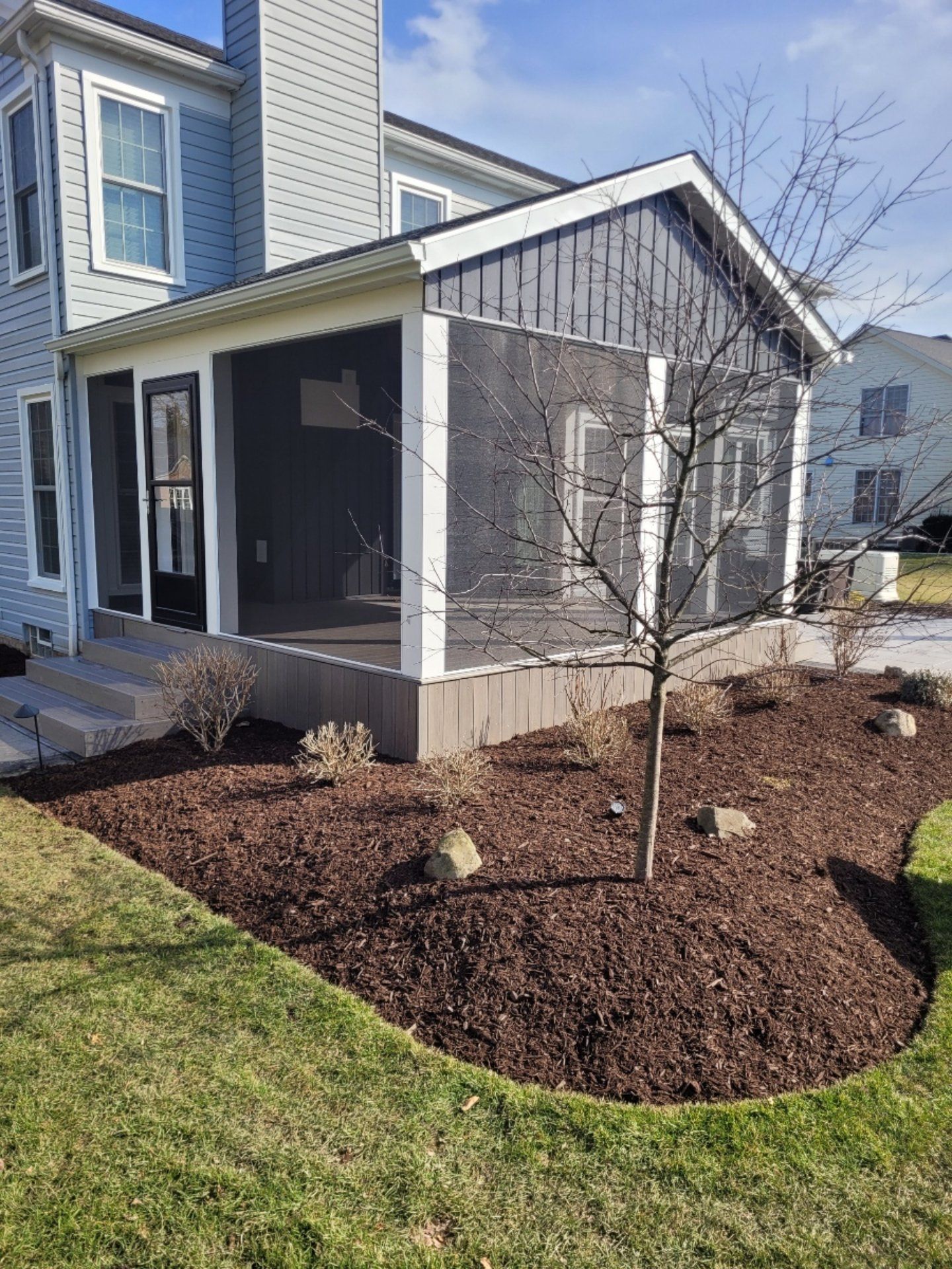 Screened porch addition to a house with dark gray siding, mulch bed, and a small tree.