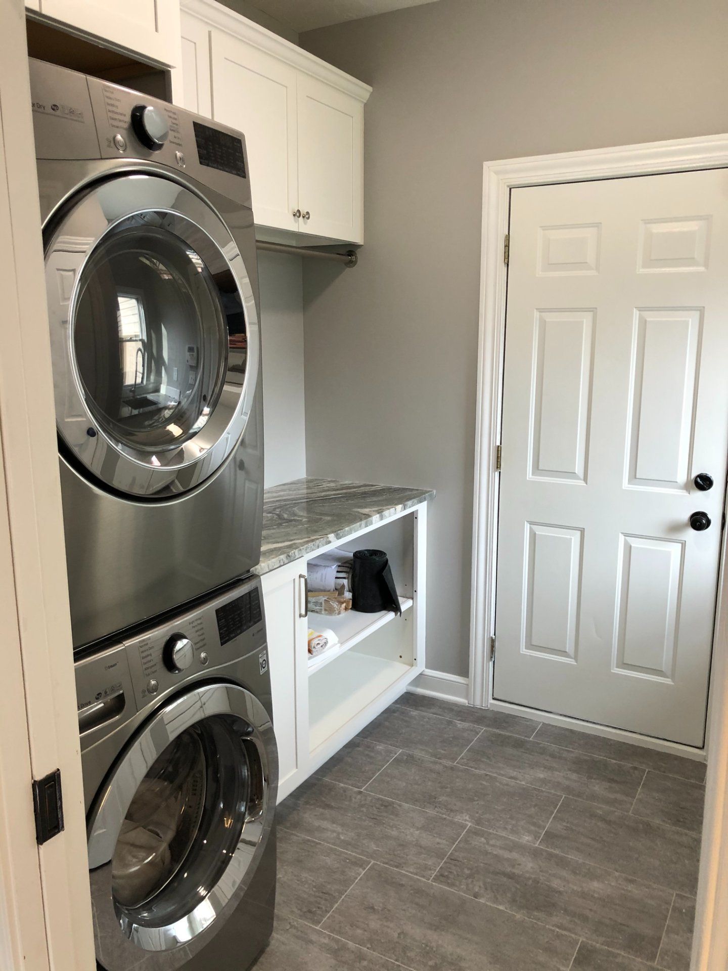 Stacked stainless steel washer and dryer in a laundry room with white cabinets, gray walls, and a door.