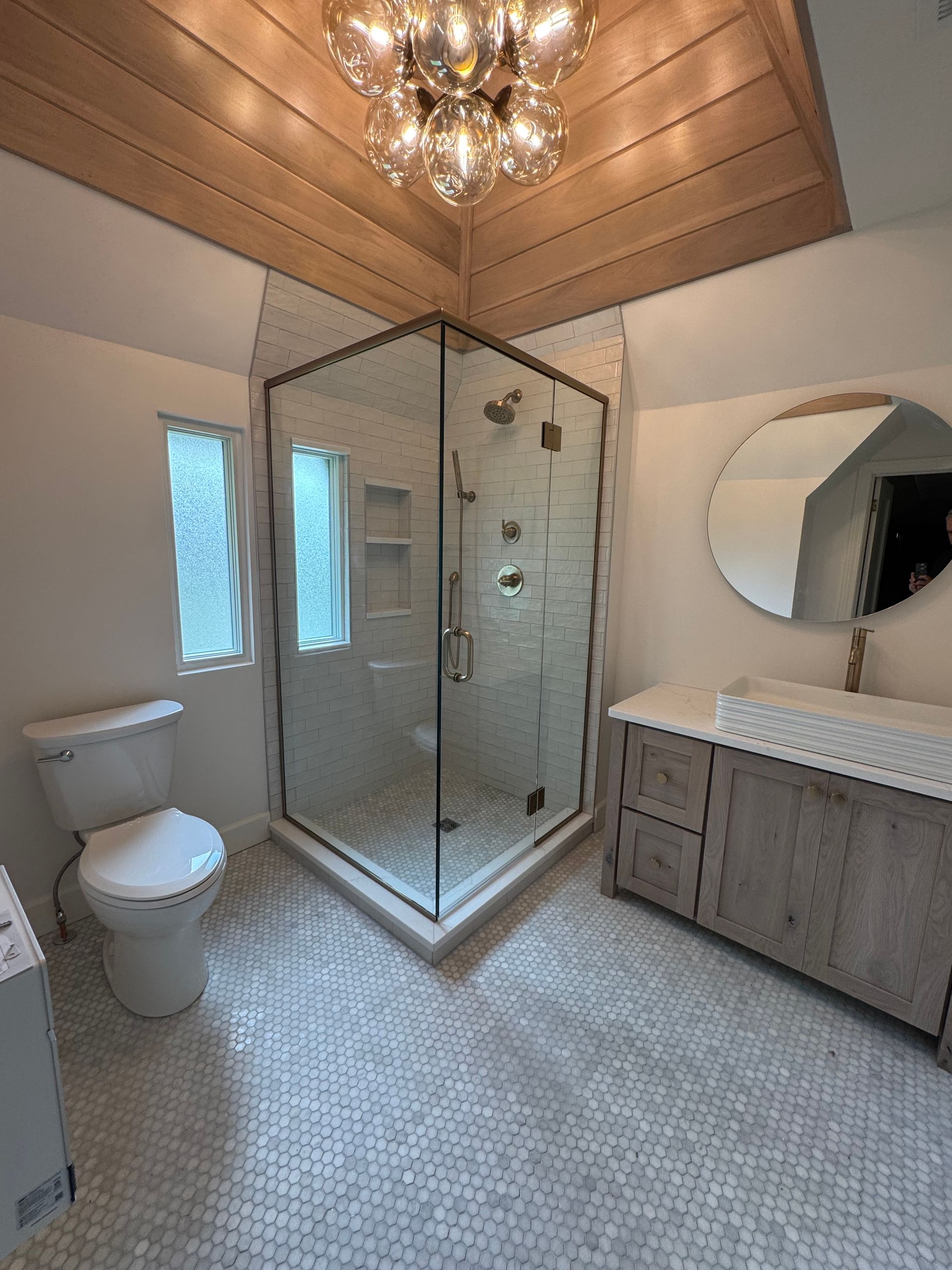 Modern bathroom with corner shower, toilet, vanity, and round mirror. White and gray tones, wood ceiling, mosaic floor.