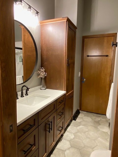 Bathroom with wooden cabinetry, white countertop, round mirror, and geometric tile floor.