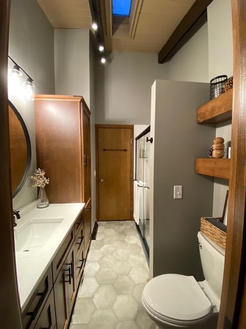 Narrow bathroom with long vanity, toilet, and shower. Gray walls, wood shelves, and hexagon tile floor.