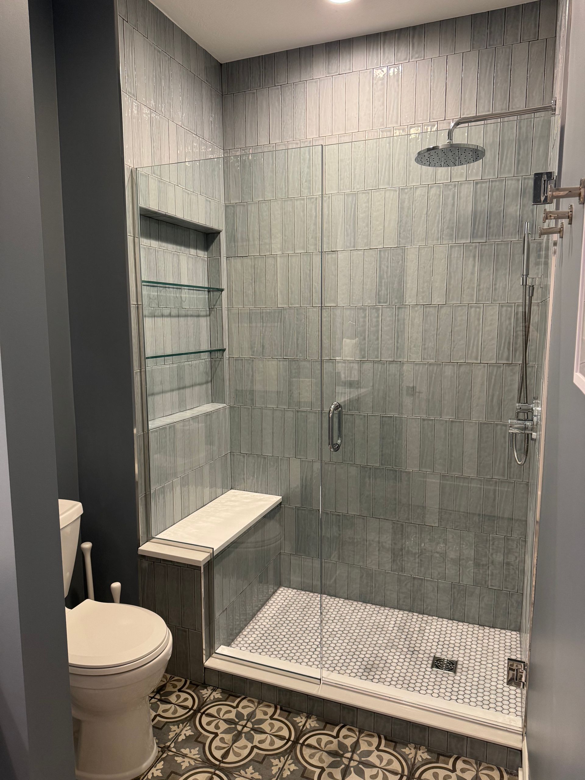 Bathroom with blue tile shower, built-in seat, and glass door. White toilet and patterned floor.