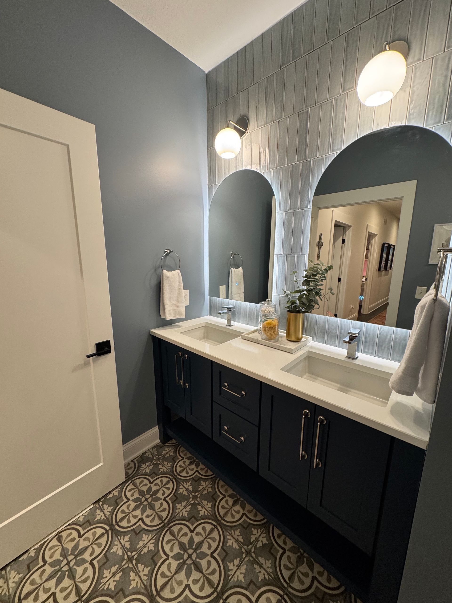 Bathroom with blue walls, patterned floor tiles, double vanity, arched mirrors, and two round sconces.