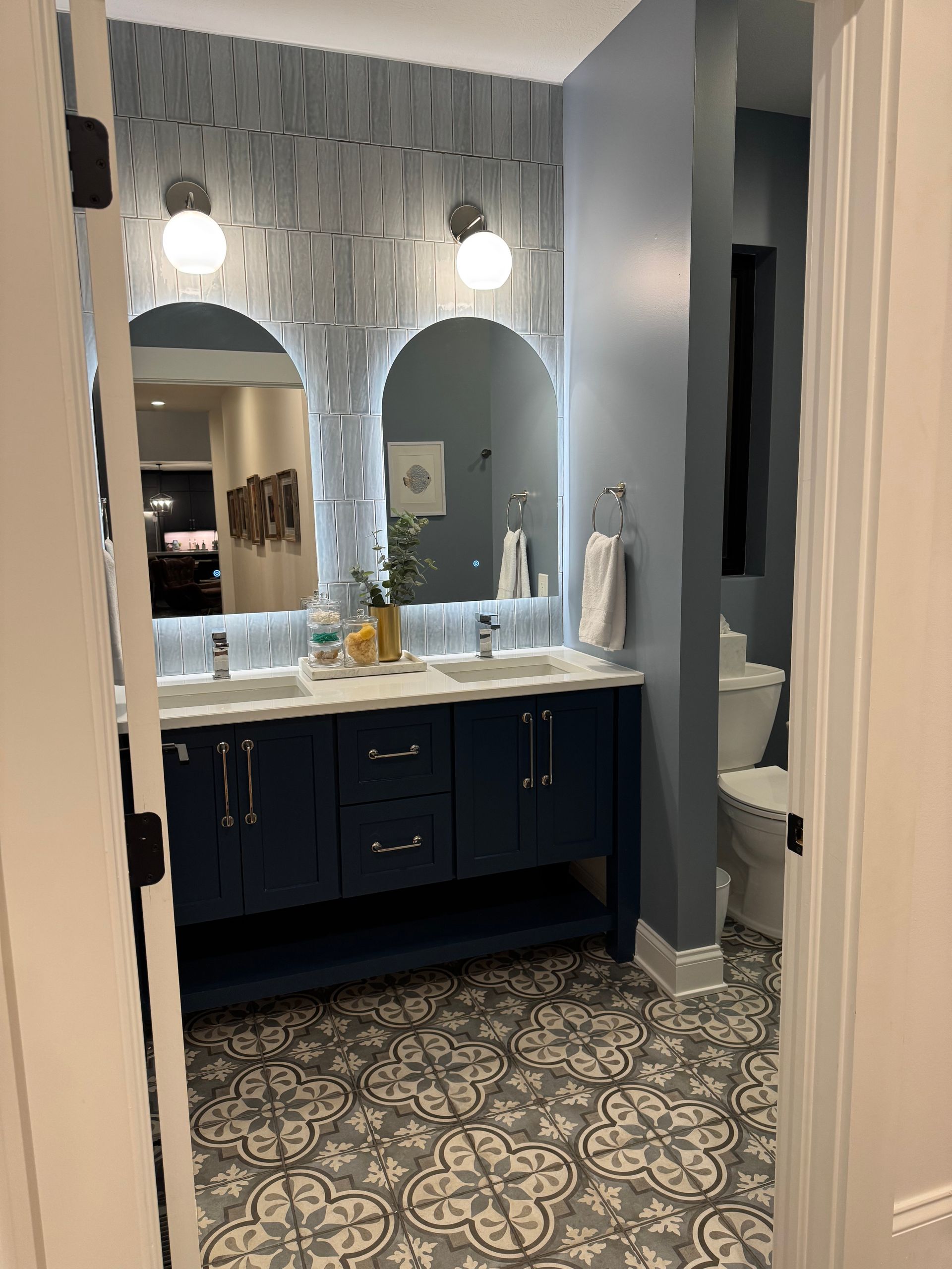 Bathroom with blue vanity, patterned tile floor, arched mirrors, and two round lights.