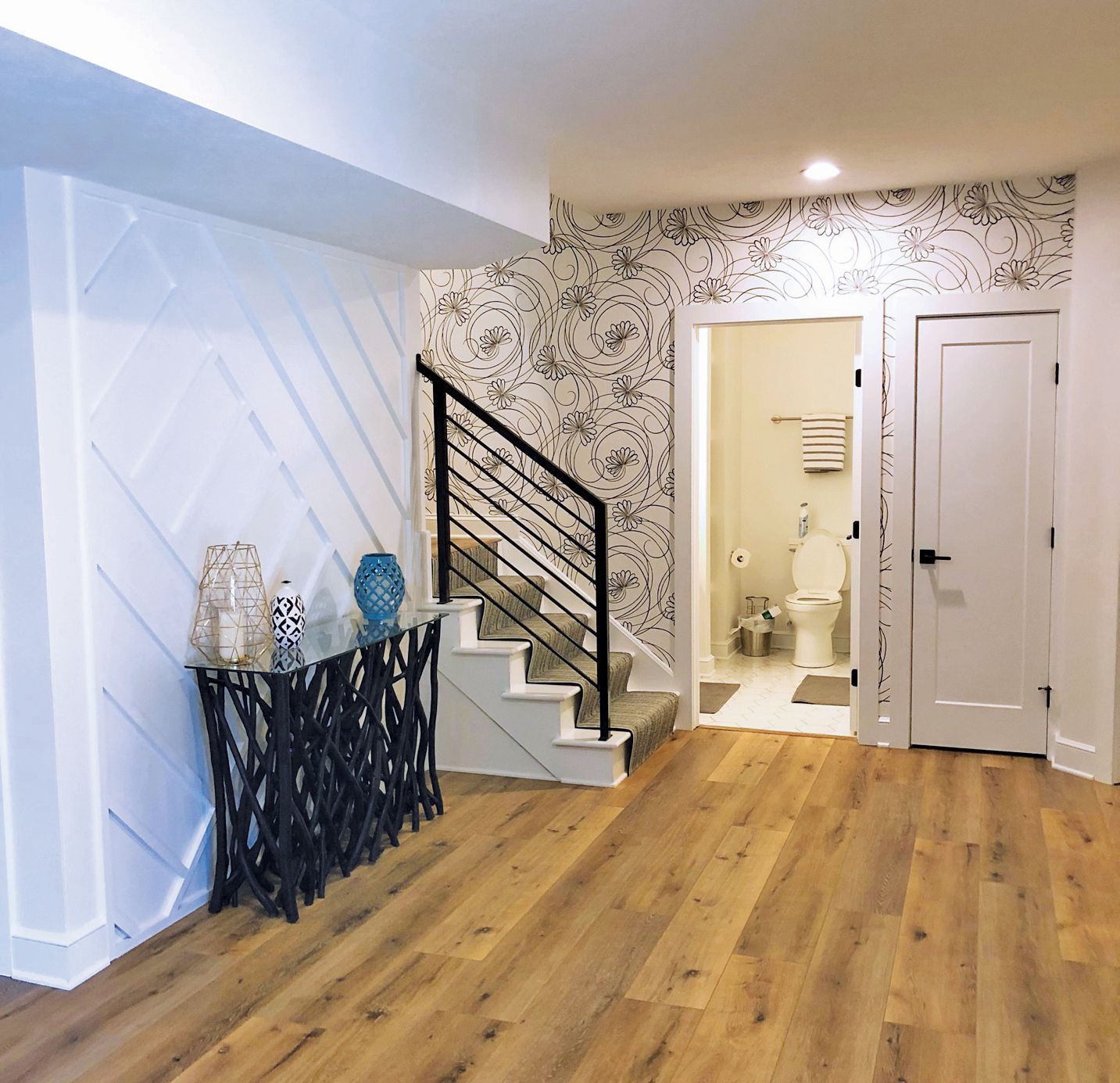 Wooden floor interior with stairs, a bathroom, and a decorative console table against a white wall with diamond paneling.