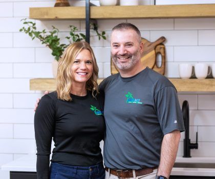 A man and woman smiling, arms around each other in a kitchen, both wearing company shirts.