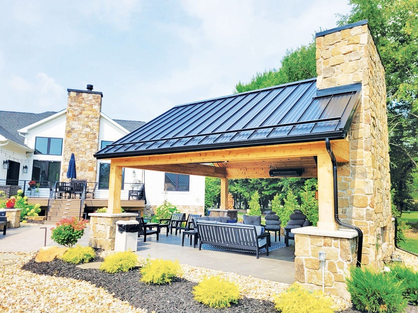Outdoor patio with stone fireplace, black metal roof, seating, and landscaping.