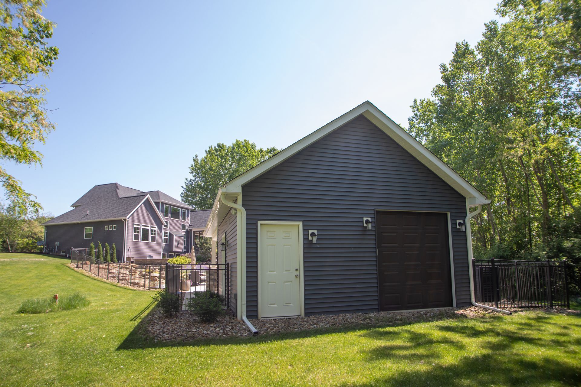 Garage with dark siding, white door, and brown garage door, in a grassy yard. House in background.