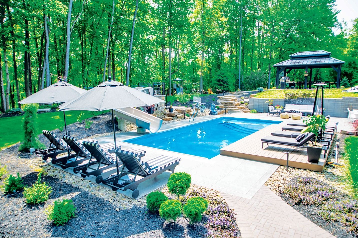 Backyard pool with lounge chairs, umbrellas, and gazebo surrounded by trees.