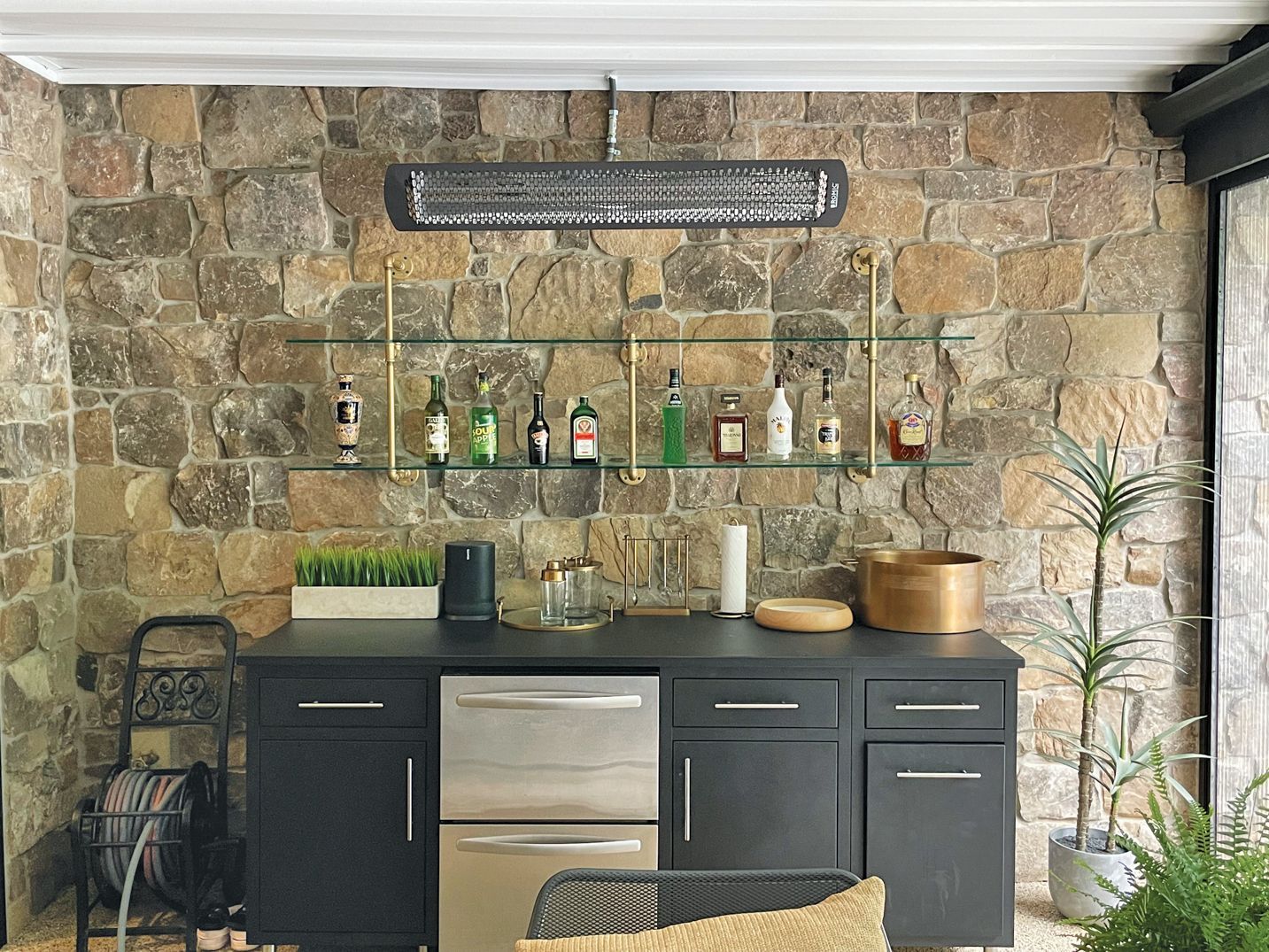 Outdoor bar with stone wall, shelves with liquor bottles, black cabinetry, and a stainless steel cooler.
