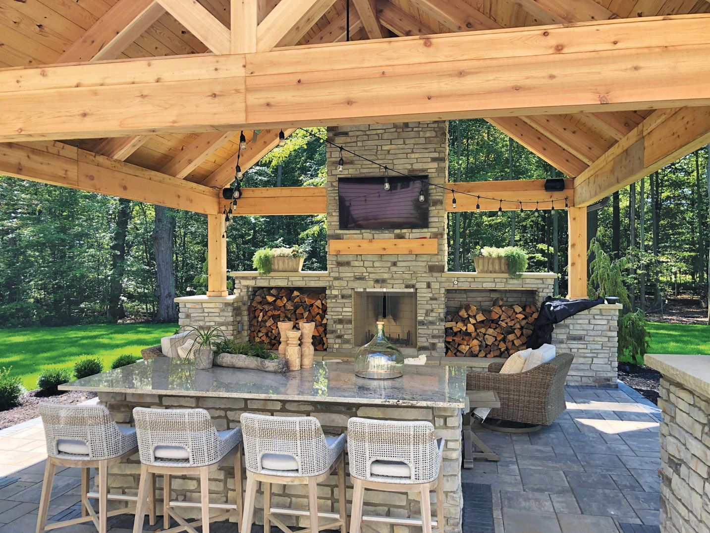 Outdoor kitchen with stone fireplace, bar, and wooden pergola. Firewood and TV are present.
