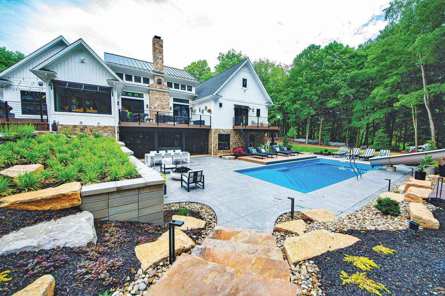 White modern farmhouse with pool, patio, and stone landscaping in a wooded setting.
