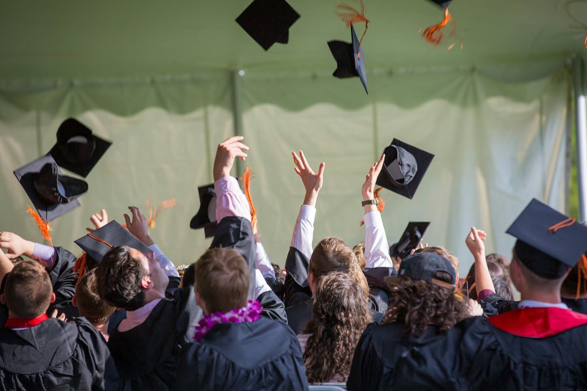 Graduates in black robes throwing caps in the air; orange tassels, green backdrop.