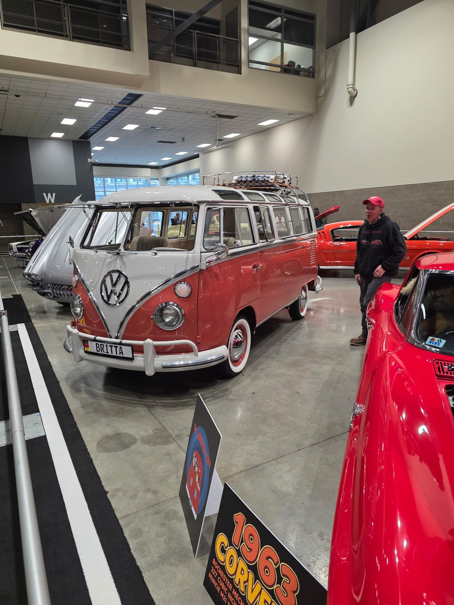 Red and white vintage VW bus with luggage rack, at an indoor car show.
