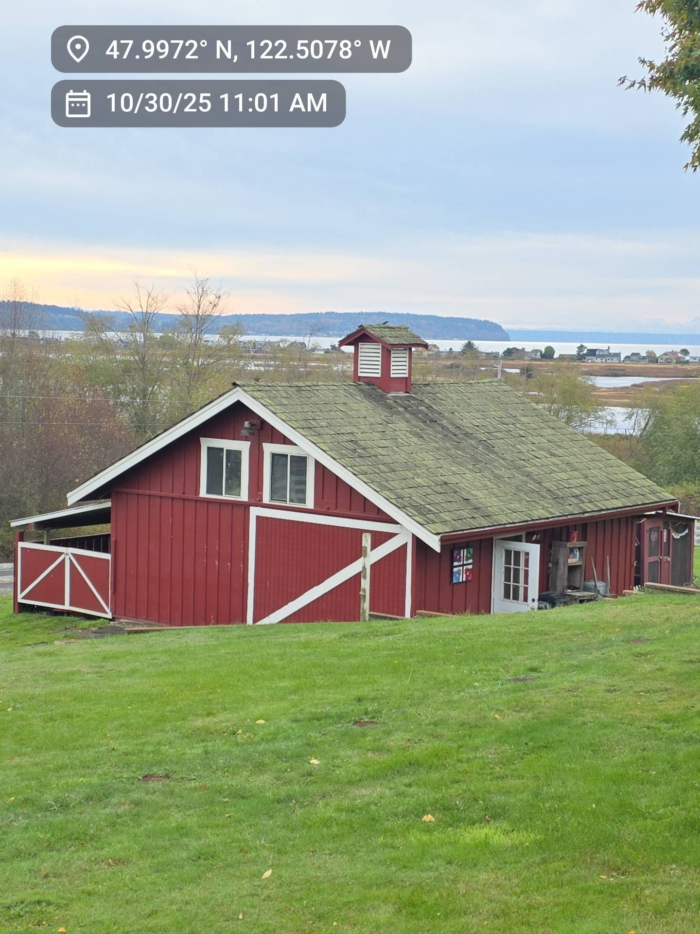 Red barn with mossy roof on a grassy hill overlooking water under a cloudy sky.