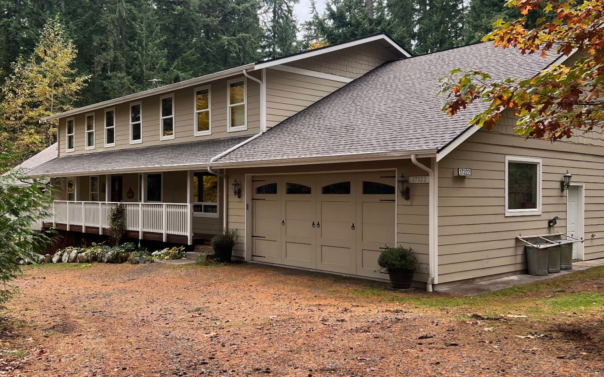 Beige two-story house with a two-car garage, porch, and a gravel driveway; set in a wooded area.