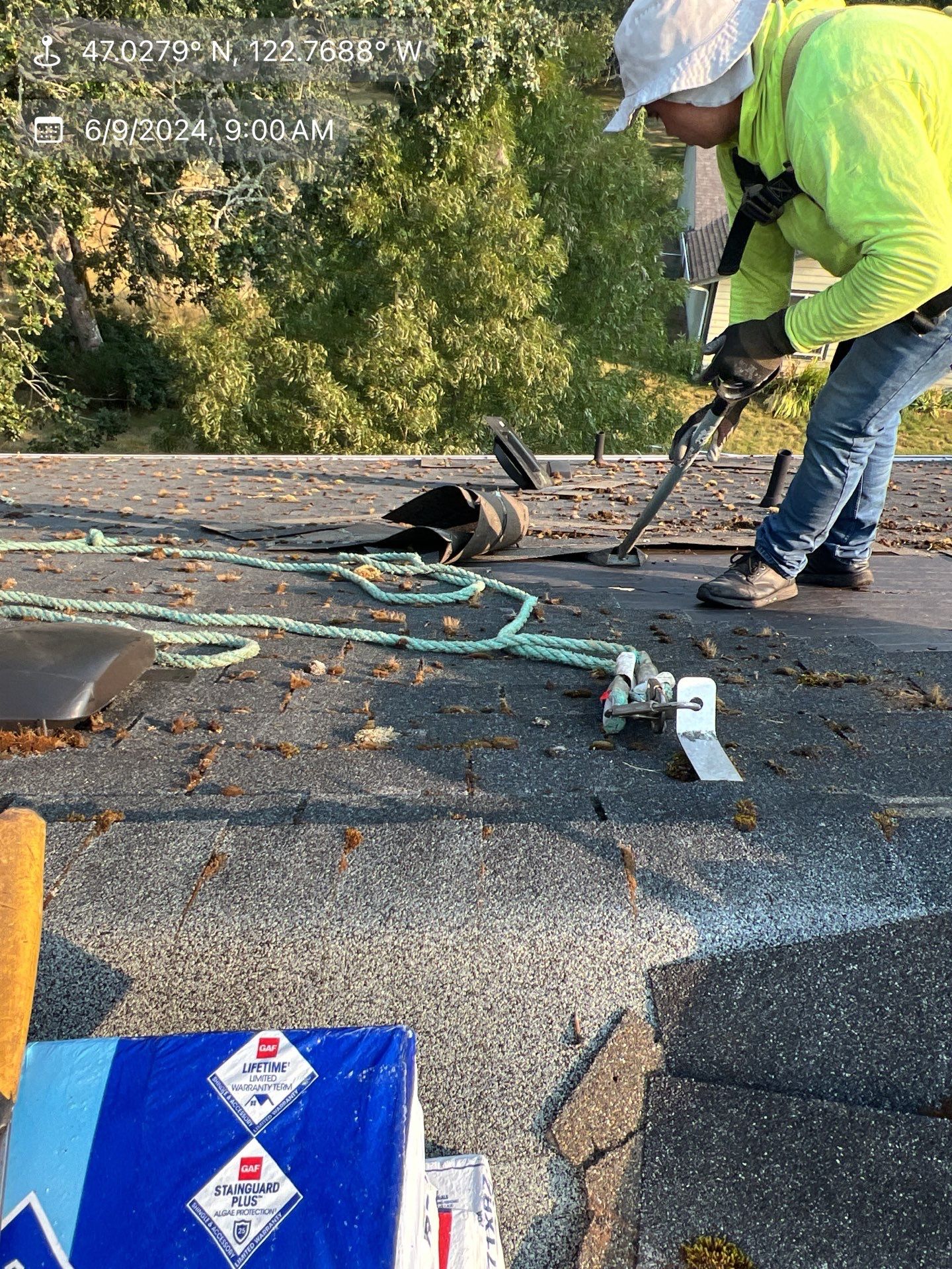Roofer on a roof, attaching safety gear. Green rope and vest, blue sky, trees in the background.