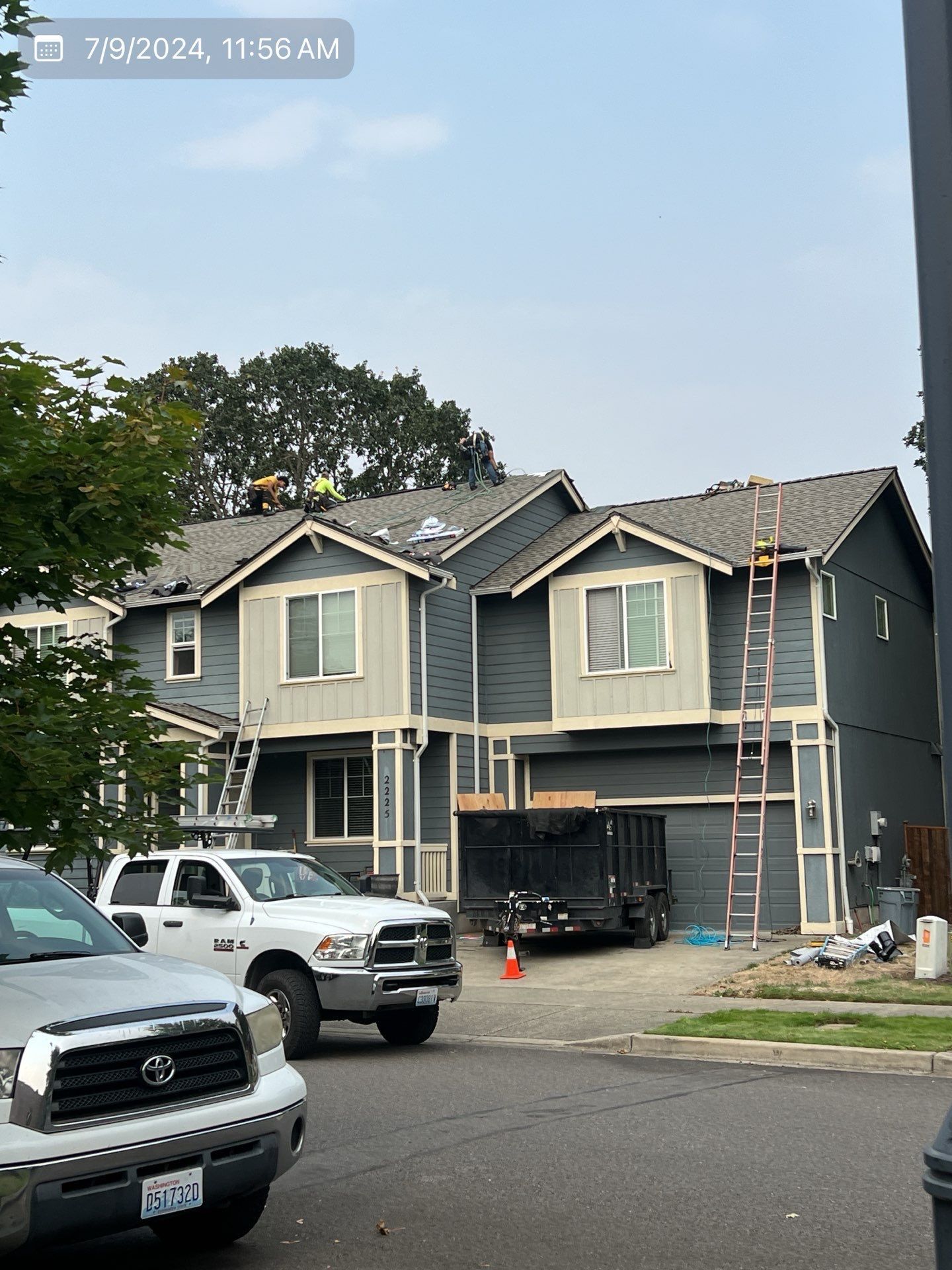 Construction workers on a two-story blue house roof, with white trucks and a black dumpster.