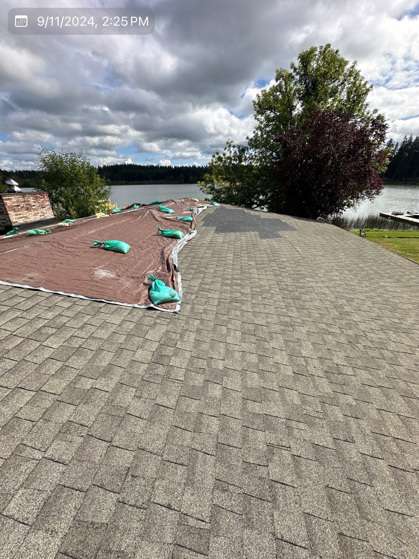 Roof with damaged shingles, some removed. Green water-filled bags on exposed area. Lake and trees in background.