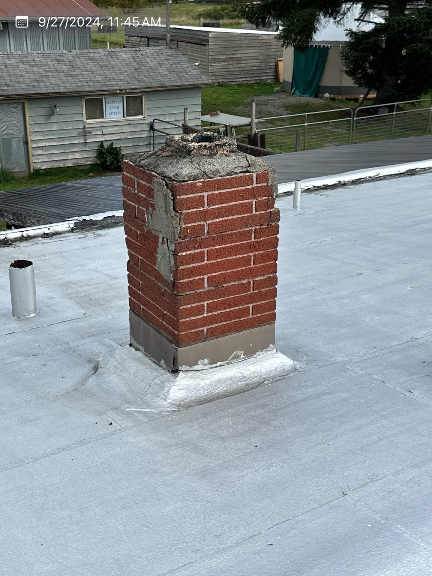 Red brick chimney on a gray roof, with signs of deterioration. In an outdoor setting.
