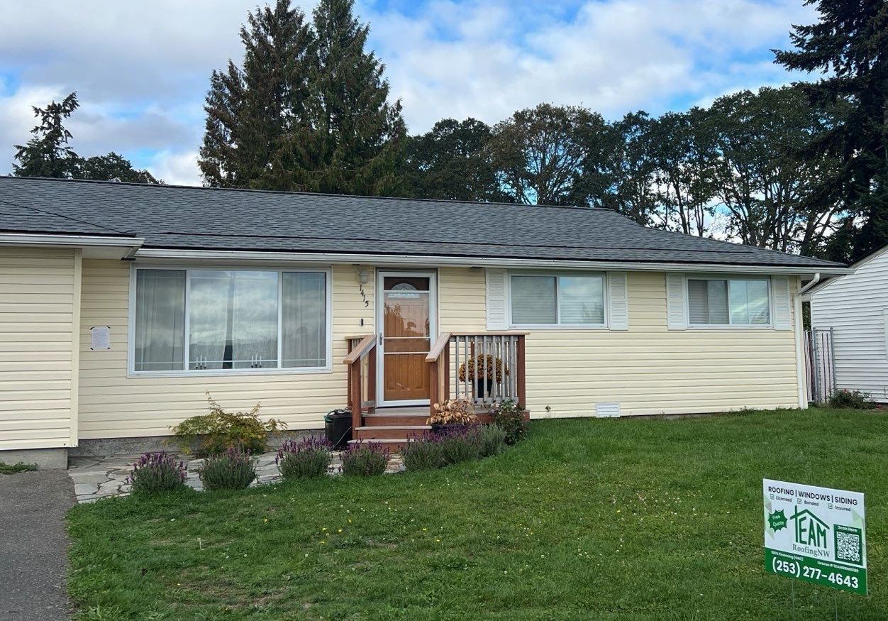 Yellow house with a dark roof and small porch, surrounded by green grass and plants.