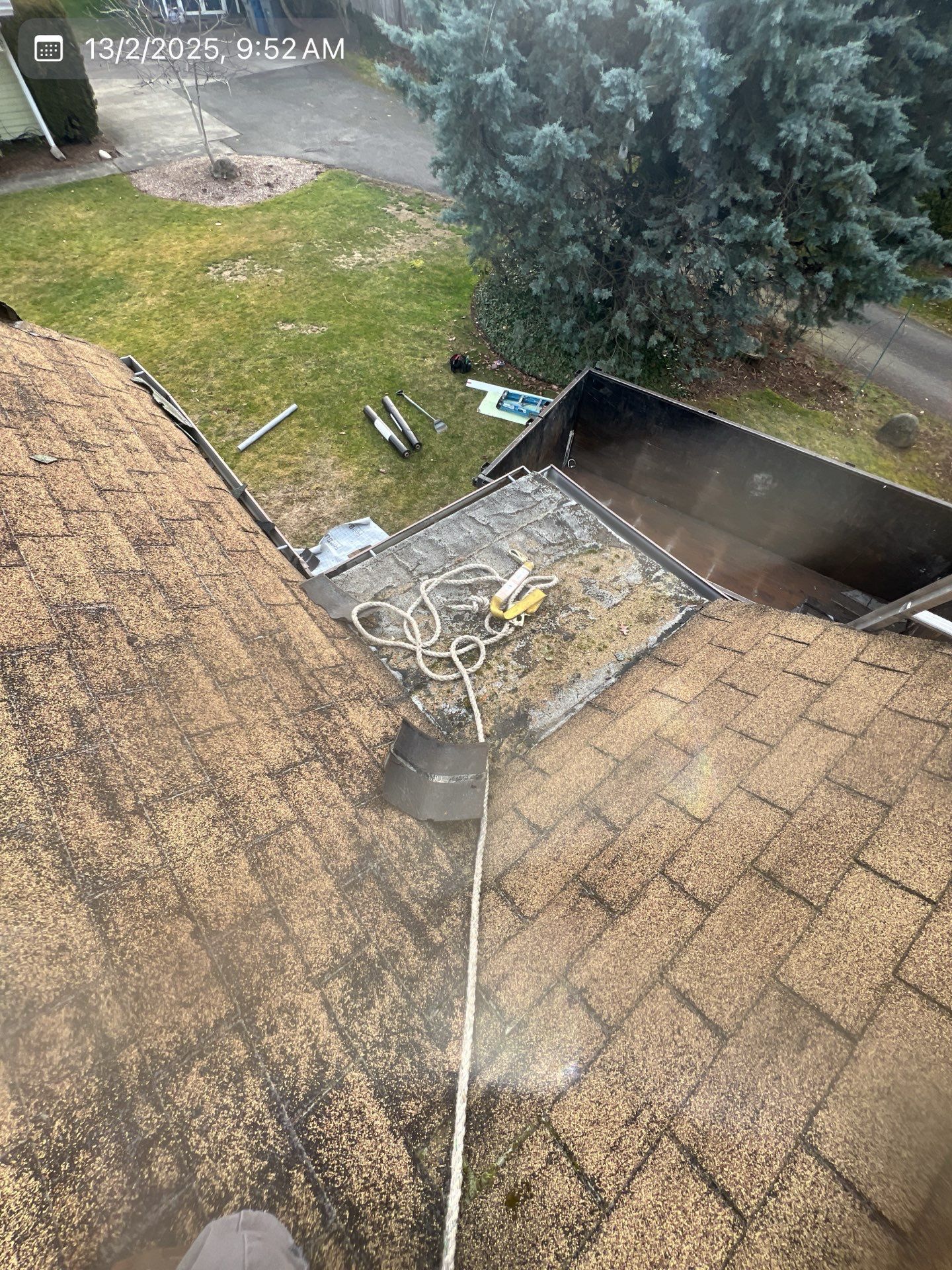 Roof being pressure washed, tools visible, person's foot, green grass, trees, and gray driveway in the background.