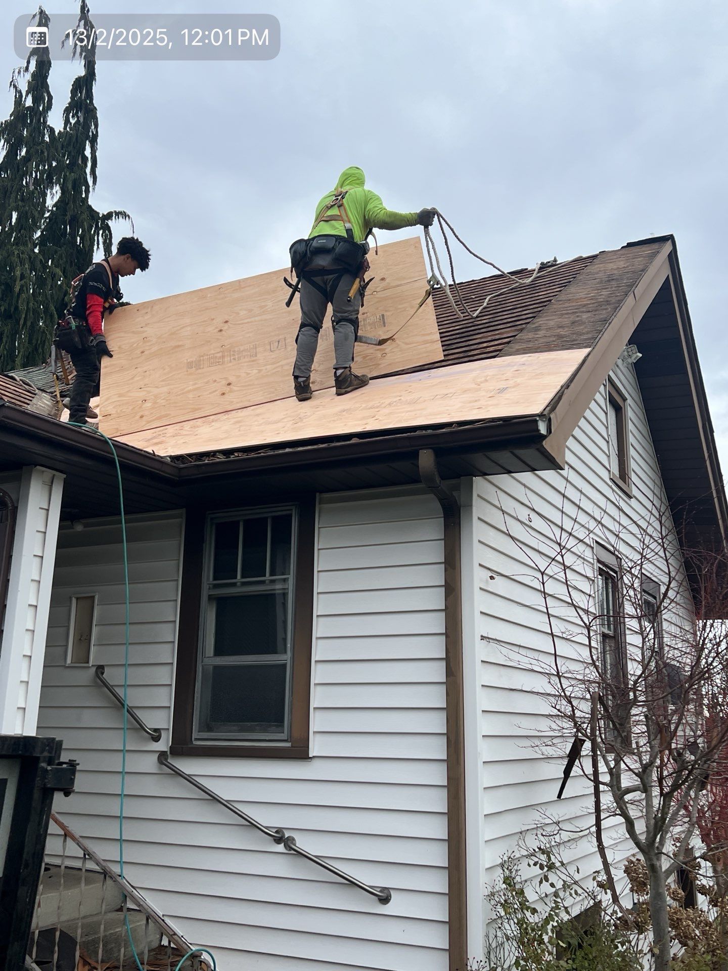 Two roofers on a house roof. One in yellow, the other in black, installing plywood underlayment. Overcast sky.