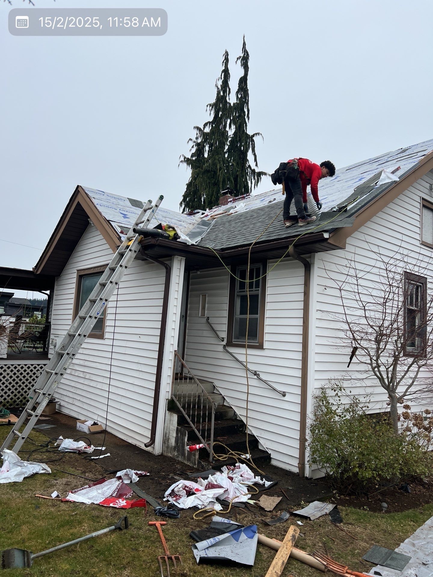 Person on a roof replacing shingles, ladder propped against the house. Debris and tools on the ground.