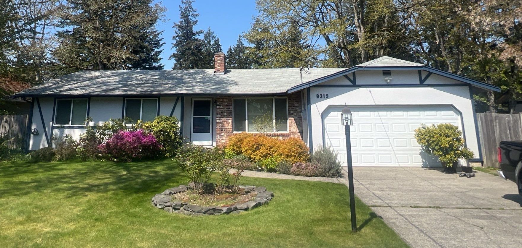 A one-story house with a white garage door, garden, and green lawn on a sunny day by Team Roofing NW in Lakewood, WA.