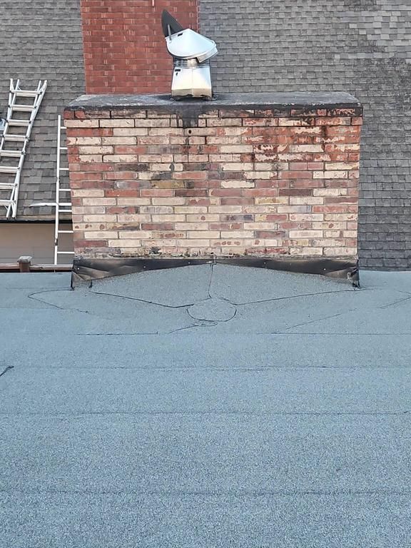 Brick chimney on a flat roof with an antenna; weathered brick, gray roof with cracks, ladder visible.